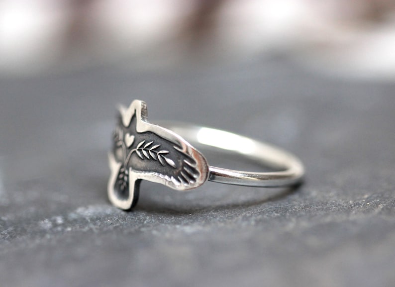 May include: A silver ring featuring a detailed bird design. The bird's head is visible, with a heart and leaf details. The ring is set against a gray background, showcasing the intricate craftsmanship of the jewelry.