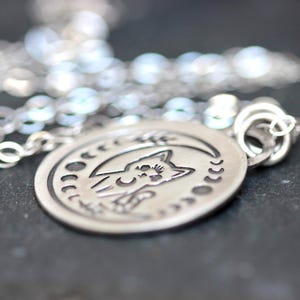 May include: A silver necklace with a round pendant featuring a cat silhouette inside a crescent moon with stars. The pendant has a detailed design and a textured finish.