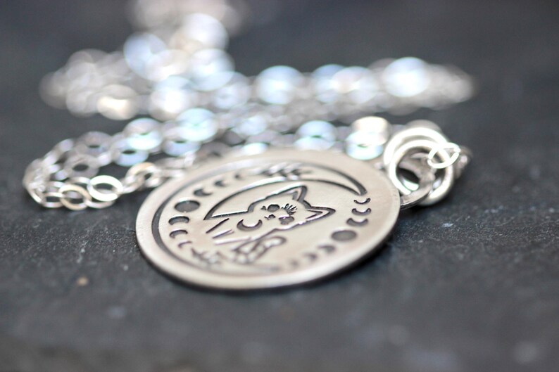 May include: A silver necklace with a round pendant featuring a cat silhouette inside a crescent moon with stars. The pendant has a detailed design and a textured finish.
