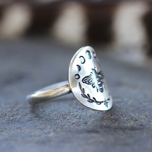 May include: A silver ring with a half-moon design featuring a bee and crescent moons. The ring is on a gray textured surface.