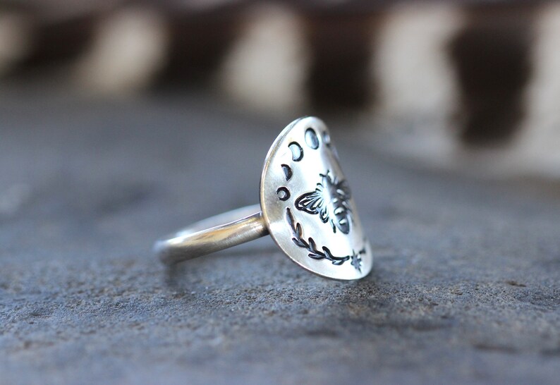 May include: A silver ring with a half-moon design featuring a bee and crescent moons. The ring is on a gray textured surface.