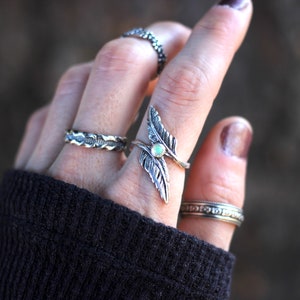 May include: A silver ring with two feather designs and a white opal stone. The ring is worn on the index finger of a hand wearing several other silver rings.
