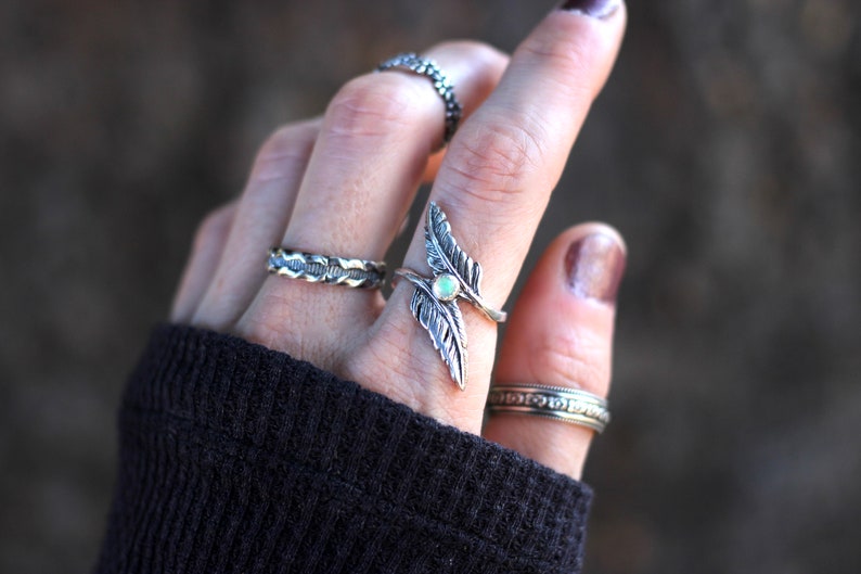 May include: A silver ring with two feather designs and a white opal stone. The ring is worn on the index finger of a hand wearing several other silver rings.