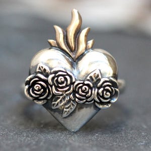 May include: A silver ring with a heart-shaped design featuring a flame and three roses. The ring is a religious symbol.
