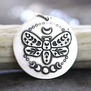 May include: A silver pendant necklace featuring a detailed, engraved design of a moth with crescent moons and stars on its wings. The pendant is round and has a textured surface.