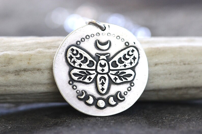 May include: A silver pendant necklace featuring a detailed, engraved design of a moth with crescent moons and stars on its wings. The pendant is round and has a textured surface.