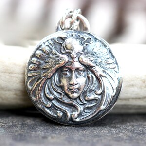 May include: A silver pendant featuring a detailed, ornate design of a woman's face with wings. The pendant is circular and has a textured, three-dimensional look.