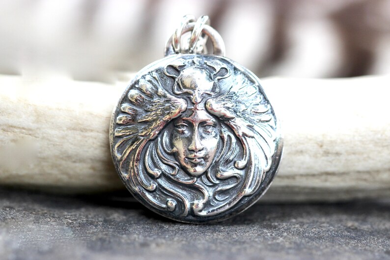May include: A silver pendant featuring a detailed, ornate design of a woman's face with wings. The pendant is circular and has a textured, three-dimensional look.