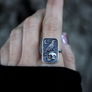 May include: A silver ring with a rectangular design featuring a raven perched on a skull. The raven is depicted in a dark gray color, while the skull is a lighter shade of gray. The ring is set against a dark background.