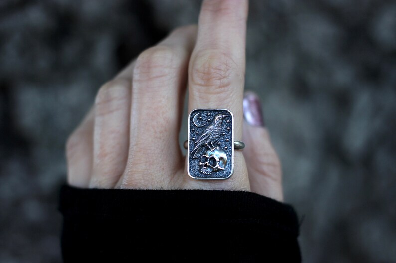 May include: A silver ring with a rectangular design featuring a raven perched on a skull. The raven is depicted in a dark gray color, while the skull is a lighter shade of gray. The ring is set against a dark background.