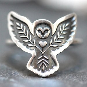 May include: A silver-toned ring featuring an owl design with outstretched wings. The owl has a heart-shaped chest and is flanked by leafy branches. The ring is detailed with etched lines and a textured finish.