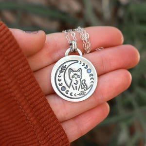 May include: Silver necklace with a crescent moon pendant featuring a cat sitting inside the moon. The pendant is surrounded by a circle of smaller moons.