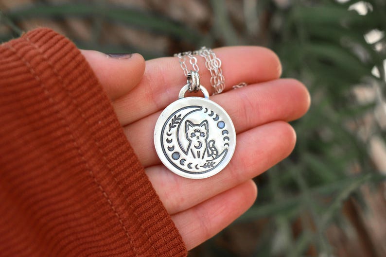 May include: Silver necklace with a crescent moon pendant featuring a cat sitting inside the moon. The pendant is surrounded by a circle of smaller moons.