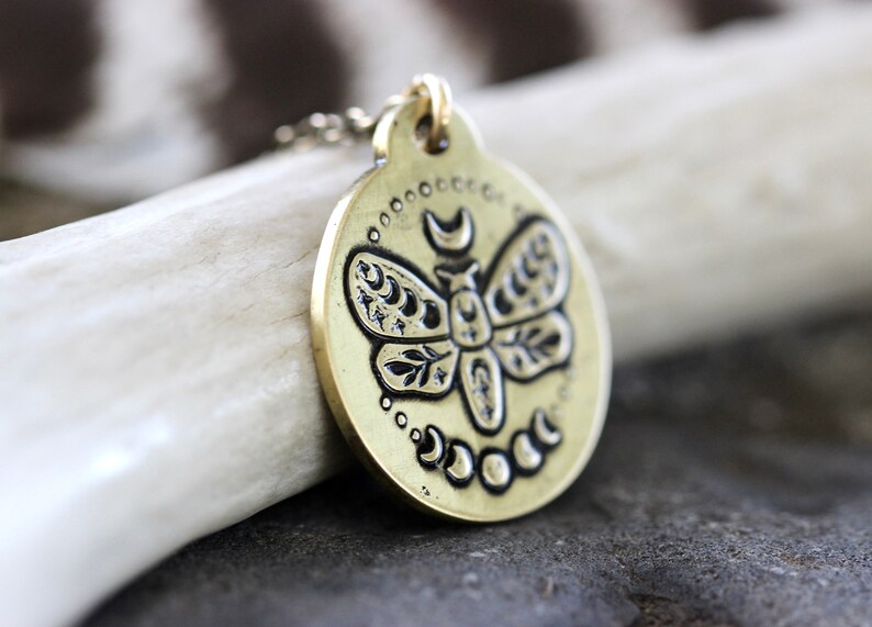 Peut inclure: Un collier en laiton avec un pendentif rond repr&eacute;sentant un motif d&eacute;taill&eacute; d'un papillon de nuit avec des croissants de lune et des &eacute;toiles sur ses ailes. Le pendentif est entour&eacute; d'un cercle de petits croissants de lune.