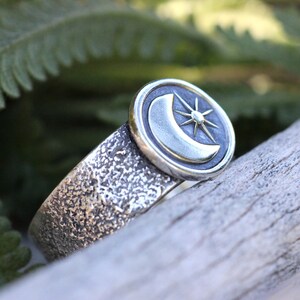 May include: A silver ring with a textured band and a circular design featuring a crescent moon and a star.