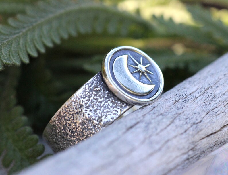 May include: A silver ring with a textured band and a circular design featuring a crescent moon and a star.