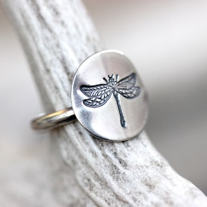 May include: A silver ring with a circular face featuring a detailed dragonfly design. The dragonfly is etched in a dark color against the polished silver. The ring is displayed on a light-colored, textured surface.