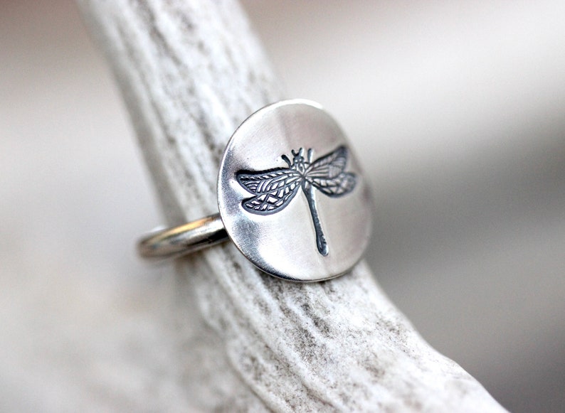May include: A silver ring with a circular face featuring a detailed dragonfly design. The dragonfly is etched in a dark color against the polished silver. The ring is displayed on a light-colored, textured surface.