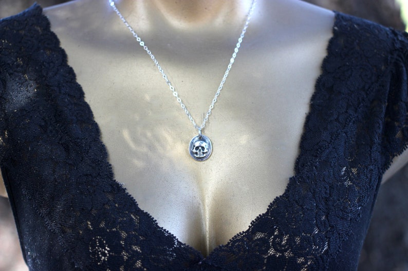 May include: A silver necklace featuring a small, detailed skull pendant. The skull is set within an oval frame and hangs from a delicate chain. The necklace is displayed against a dark, lacy garment, creating a striking contrast. The jewelry is a gothic or alternative style.