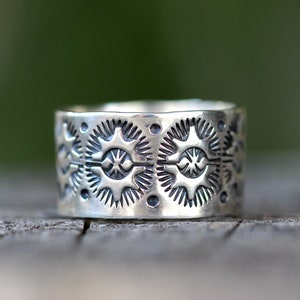 May include: A wide, silver ring with an intricate pattern of repeating starburst and dot designs. The ring's surface is textured, and the design is etched into the metal. The ring is displayed on a wooden surface.