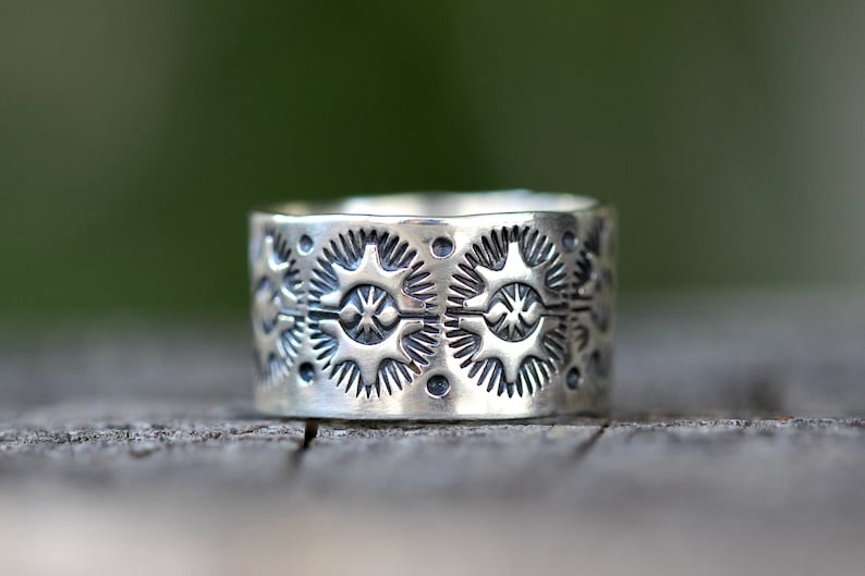May include: A wide, silver ring with an intricate pattern of repeating starburst and dot designs. The ring's surface is textured, and the design is etched into the metal. The ring is displayed on a wooden surface.