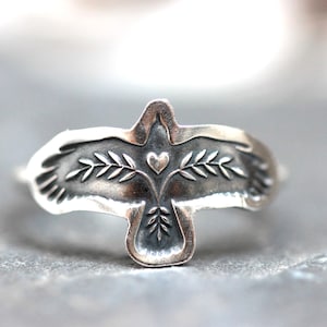 May include: A silver ring featuring a stylized bird design with outstretched wings. The bird's body includes a heart and olive branch details. The ring is made of a shiny, metallic material.