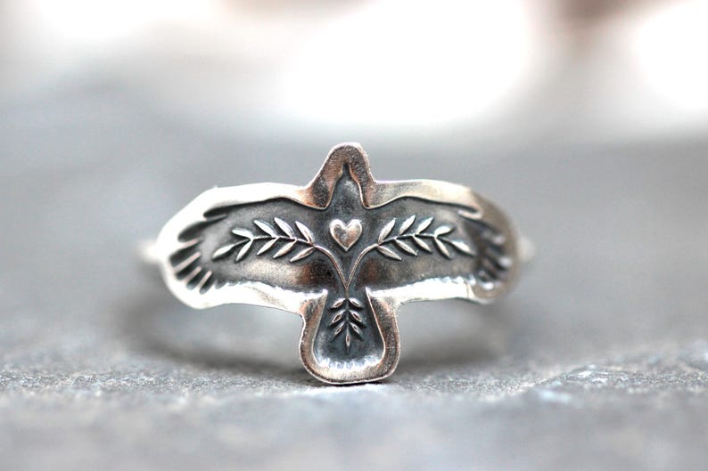 May include: A silver ring featuring a stylized bird design with outstretched wings. The bird's body includes a heart and olive branch details. The ring is made of a shiny, metallic material.