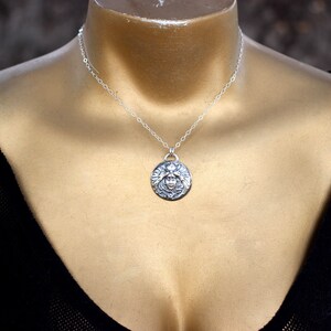 May include: A silver necklace with a round pendant featuring a detailed design of a face with wings. The necklace is on a mannequin wearing a black top.