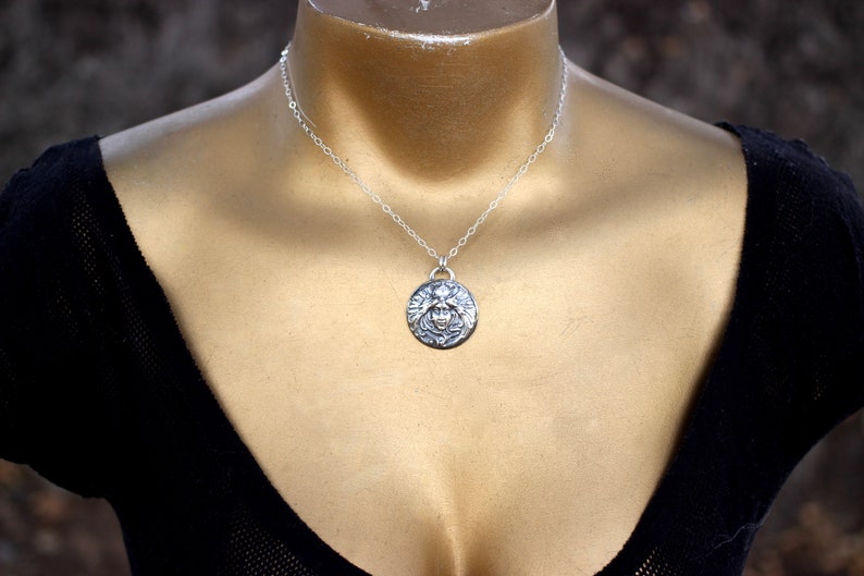 May include: A silver necklace with a round pendant featuring a detailed design of a face with wings. The necklace is on a mannequin wearing a black top.