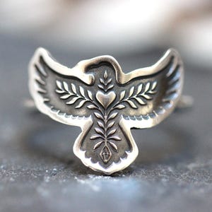 May include: A silver-toned ring featuring a stylized bird design. The bird has outstretched wings and a detailed body with leaf and heart motifs. The ring is set against a blurred, dark background, highlighting the intricate metalwork.