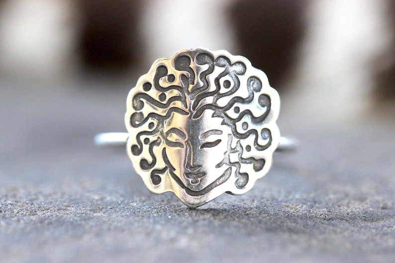 May include: A silver ring with a detailed face design and flowing hair. The face has closed eyes and a serene expression. The ring's design is circular with an ornate border, and the band is a simple, thin silver. The ring is on a grey surface.