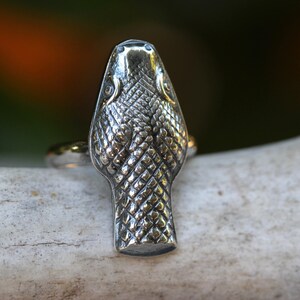 May include: A silver snake head ring with a detailed textured design. The ring is on a white background.