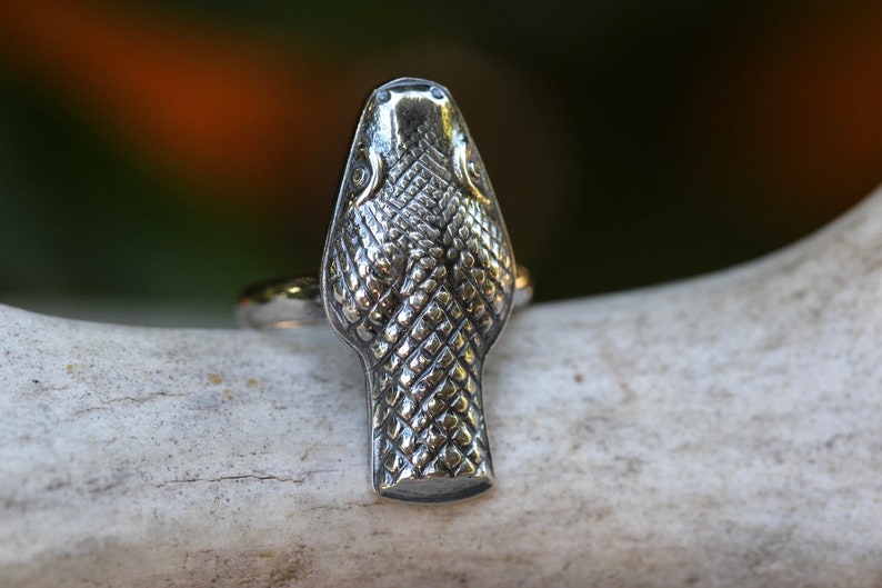 May include: A silver snake head ring with a detailed textured design. The ring is on a white background.