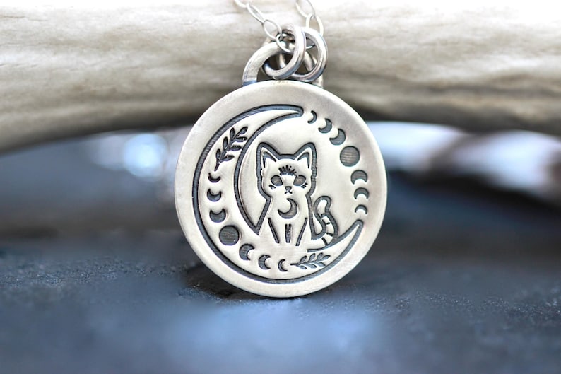 May include: A silver pendant necklace featuring a cat sitting on a crescent moon with a moon phase design. The pendant is round and has a detailed design.