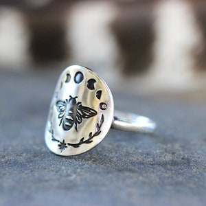 May include: A silver ring with a bee and moon phases design. The ring is oval shaped and has a textured surface.