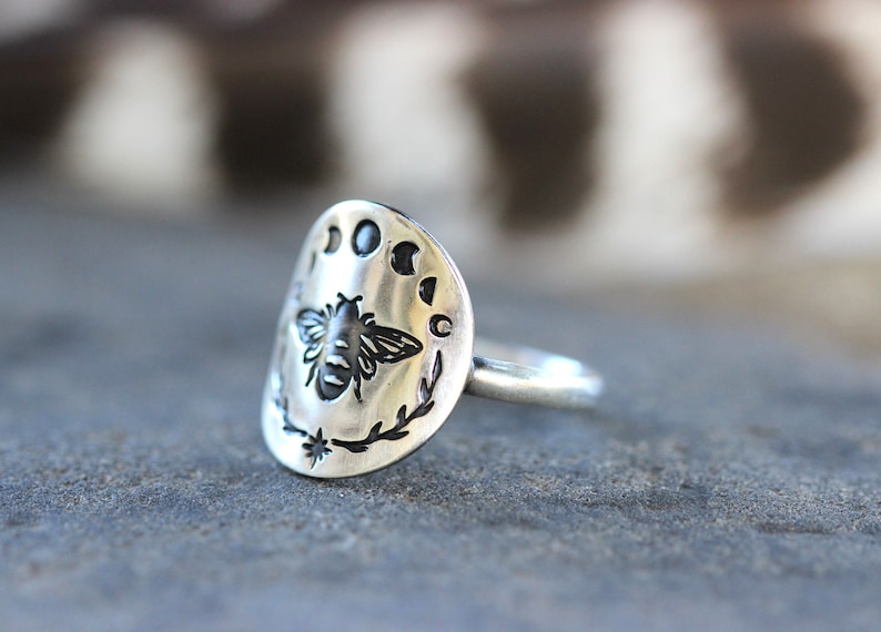 May include: A silver ring with a bee and moon phases design. The ring is oval shaped and has a textured surface.