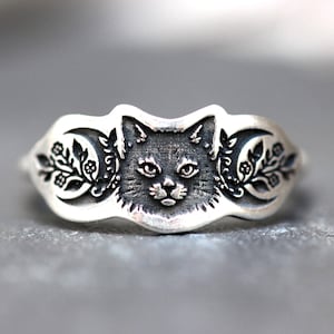 May include: A silver-toned ring featuring a detailed cat face in the center. The ring has floral and crescent moon designs on either side of the cat. The ring is likely made of sterling silver.