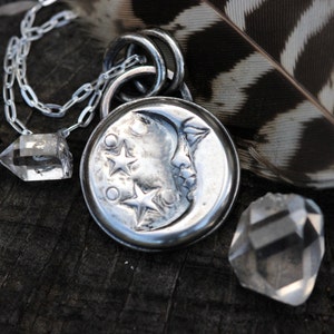 Puede incluir: Un collar de plata con un colgante de luna creciente con tres estrellas. El colgante est&aacute; unido a una cadena de plata y est&aacute; acompa&ntilde;ado por dos cristales de cuarzo transparentes.