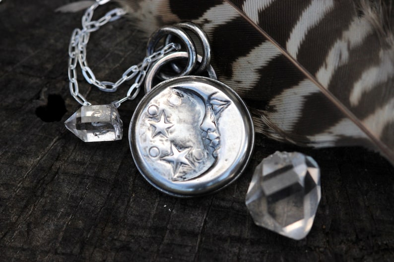 Puede incluir: Un collar de plata con un colgante de luna creciente con tres estrellas. El colgante est&aacute; unido a una cadena de plata y est&aacute; acompa&ntilde;ado por dos cristales de cuarzo transparentes.