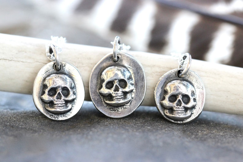 May include: Three silver-toned pendants, each featuring a detailed skull design. The pendants are oval and circular, suspended from delicate chains. The skull's features are clearly defined, creating a gothic or edgy aesthetic.