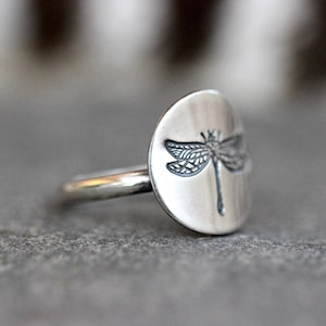 May include: A silver ring featuring a dragonfly design. The ring has a round, flat face with an engraved dragonfly. The band is thin and simple. The ring is set against a gray background.
