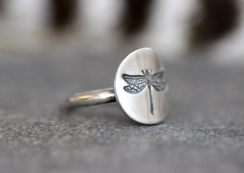 May include: A silver ring featuring a dragonfly design. The ring has a round, flat face with an engraved dragonfly. The band is thin and simple. The ring is set against a gray background.
