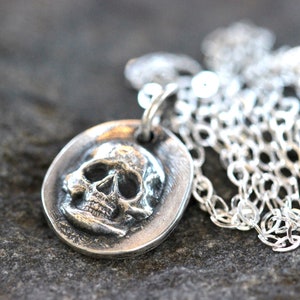 May include: A silver-toned pendant necklace featuring a detailed skull design. The skull is set within an oval frame and hangs from a delicate chain. The necklace is displayed against a textured, gray background.