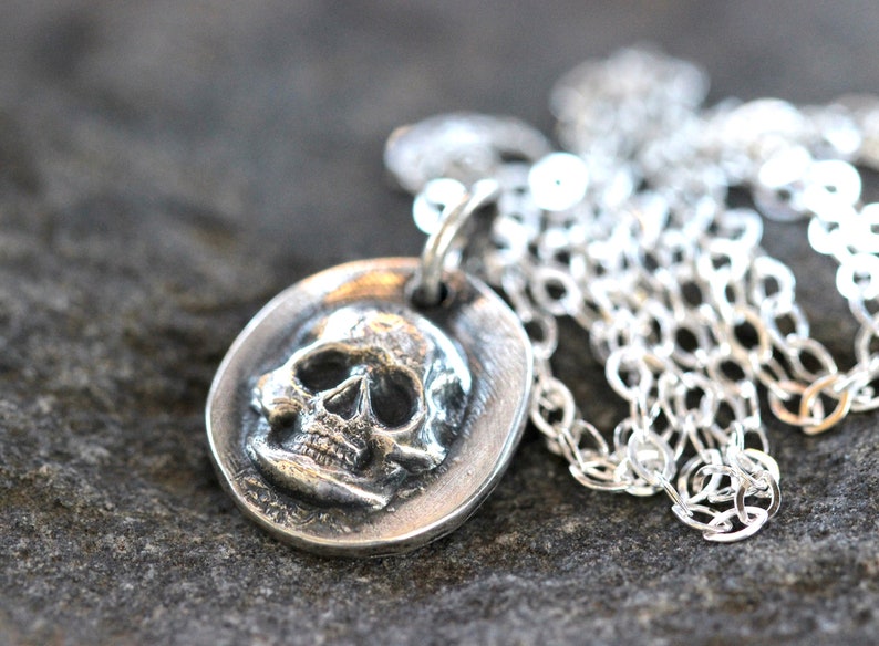 May include: A silver-toned pendant necklace featuring a detailed skull design. The skull is set within an oval frame and hangs from a delicate chain. The necklace is displayed against a textured, gray background.