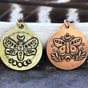 Peut inclure: Deux pendentifs ronds en m&eacute;tal avec un motif d&eacute;taill&eacute; d'un papillon de nuit avec des accents de lune et d'&eacute;toiles. Le pendentif de gauche est en laiton et le pendentif de droite est en cuivre.