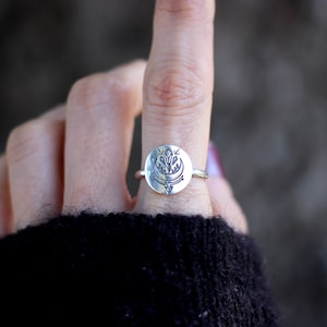 May include: A silver ring with a circular design featuring a stylized floral motif. The ring is worn on a finger.