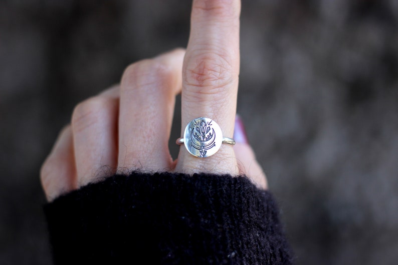 May include: A silver ring with a circular design featuring a stylized floral motif. The ring is worn on a finger.