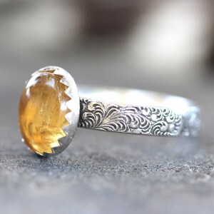 May include: A silver ring with an oval-shaped, yellow gemstone set in a scalloped bezel. The band is decorated with an intricate floral design.