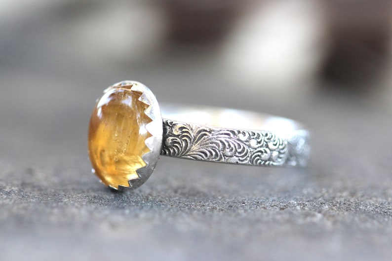 May include: A silver ring with an oval-shaped, yellow gemstone set in a scalloped bezel. The band is decorated with an intricate floral design.