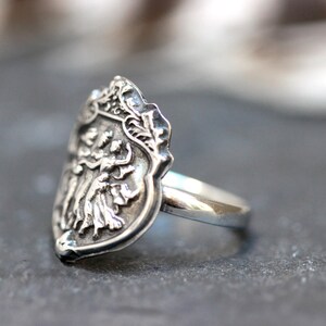 Puede incluir: Un anillo de plata con un dise&ntilde;o de escudo de armas detallado que presenta tres figuras. El anillo est&aacute; hecho de plata de ley y tiene un acabado pulido.